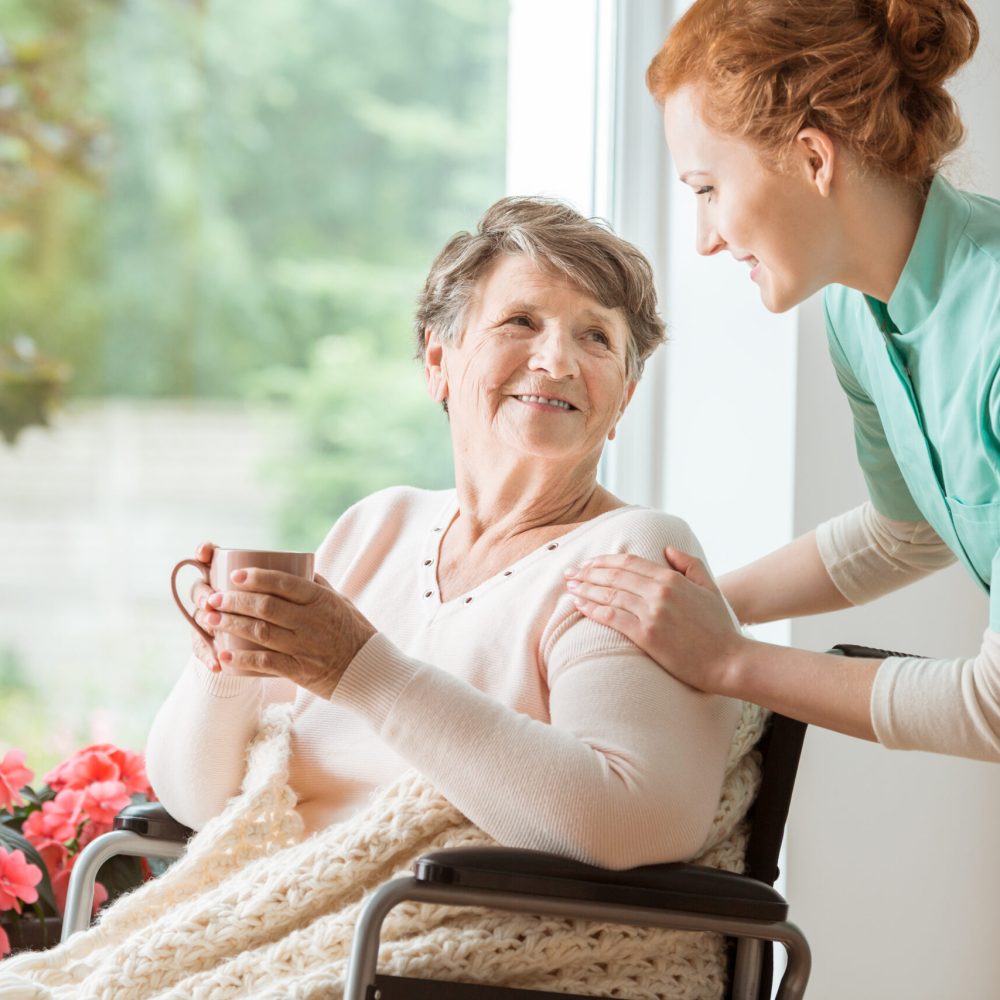 A professional caretaker in uniform helping a geriatric female patient on a wheelchair. Senior holding a cup and sitting by a large window in a rehabilitation center.